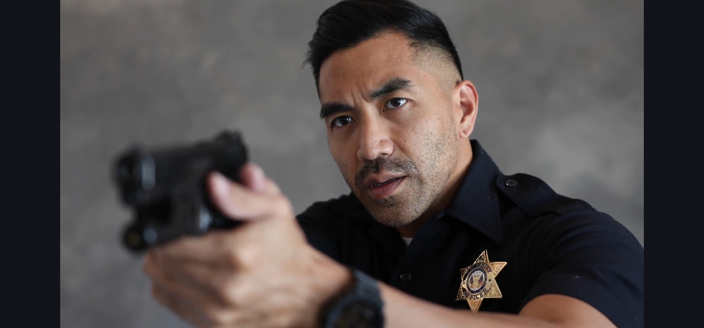 An actor performing a self-tape as a police officer, aiming a prop gun at the camera